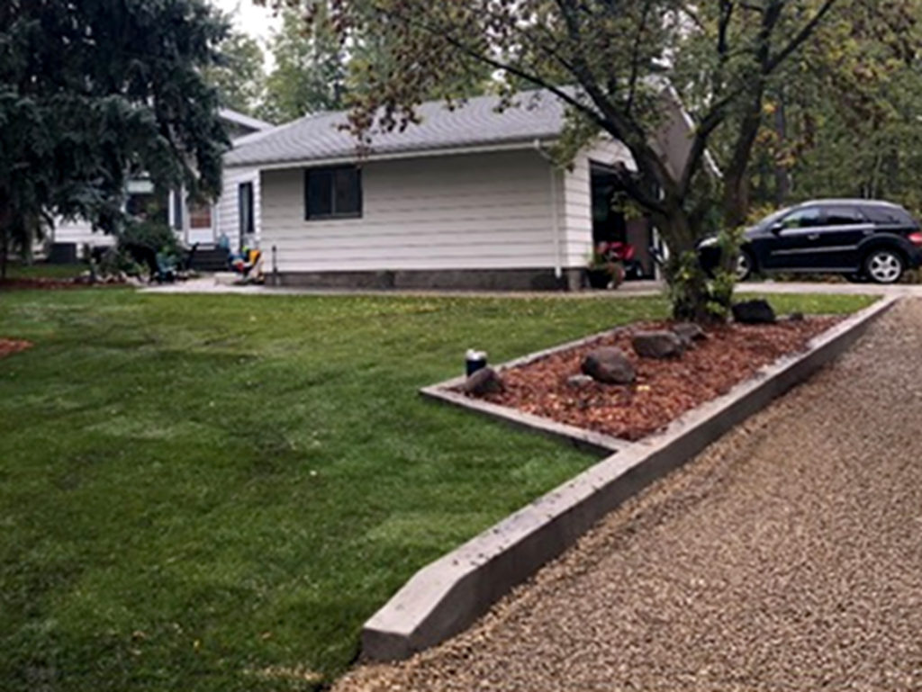 Residential yard with concrete curbs/edging around trees and well area, and gravelled driveway. As well as landscaping (sod, rocks, and wood chips) for back acreage yard.